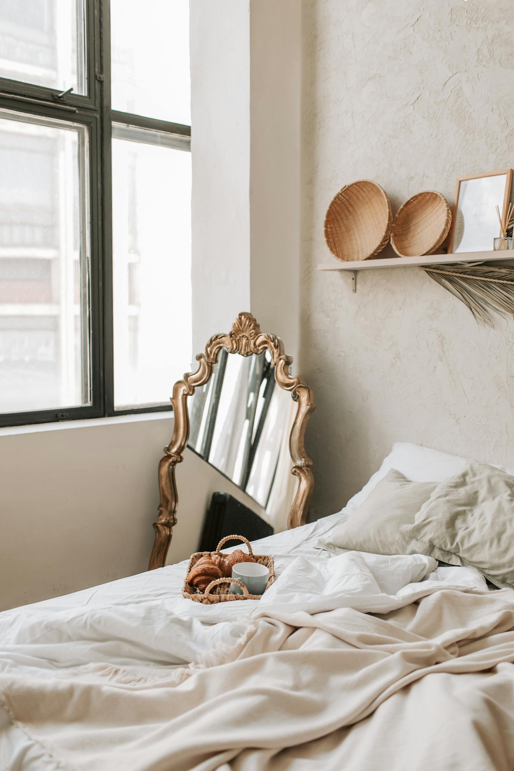 Cozy bedroom scene featuring a vintage mirror, pillows, and a basket on a soft bed by a bright window.